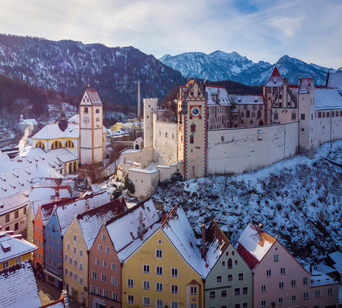 Das Bild fängt die atemberaubende Sicht auf das mittelalterliche Städtchen Füssen in Deutschland ein. Die Stadt liegt auf einem Hügel, von majestätischen Bergen umgeben. Die Architektur der Stadt ist durch bunte Gebäude geprägt, jedes mit einem einzigartigen Charme. Die Gebäude sind mit Schnee verziert, was eine Prise Winter in die Szene bringt