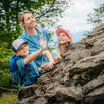 Das Bild zeigt eine Frau und zwei Kinder auf einer felsigen Oberfläche, mit der Frau, die nach oben zeigt. Die Frau lächelt, und die Kinder schauen zu ihr hinauf.