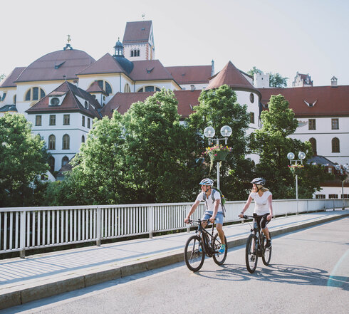Das Bild zeigt einen Mann und eine Frau beim Fahrradfahren. Sie fahren auf E-Bikes, tragen Freizeitkleidung und Helme. Sie fahren auf den Betrachter zu auf einer asphaltierten Brücke. Im Hintergrund ist der umfangreiche Gebäudekomplex des ehemaligen Benediktinerklosters St. Mang zu sehen. Es hat eine weiße Fassade, unzählige Fenster und rote Dächer.  Die Sonne scheint.