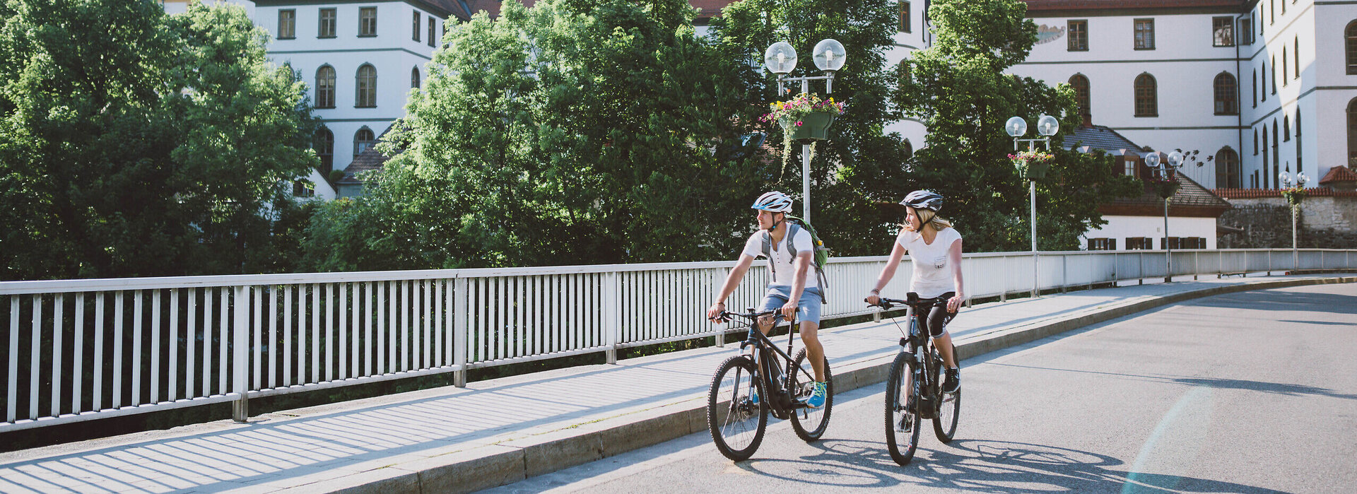 Das Bild zeigt einen Mann und eine Frau beim Fahrradfahren. Sie fahren auf E-Bikes, tragen Freizeitkleidung und Helme. Sie fahren auf den Betrachter zu auf einer asphaltierten Brücke. Im Hintergrund ist der umfangreiche Gebäudekomplex des ehemaligen Benediktinerklosters St. Mang zu sehen. Es hat eine weiße Fassade, unzählige Fenster und rote Dächer.  Die Sonne scheint.