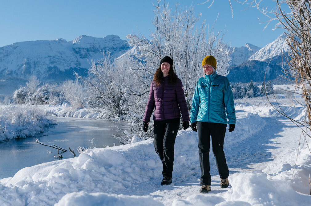 Das Bild zeigt eine friedliche Winterlandschaft. Zwei Frauen in Winterklamotten sind auf einem mit Schnee bedeckten Pfad zu sehen. Die Person auf der linken Seite trägt eine lilafarbene Winterjacke, während diejenige auf der rechten Seite eine blaue Winterjacke trägt. Der Weg, den sie entlanglaufen, ist mit Schnee bedeckt, nur ihre Fußabdrücke sind sichtbar. Der Hintergrund des Bildes ist ein atemberaubender Blick auf schneebedeckte Berge, was zur Ruhe der Szene beiträgt. Der Himmel ist klar und blau, frei von Wolken. Das Gesamtbild zeichnet ein Bild eines friedlichen Wintertages, mit den beiden Personen, die ihren Spaziergang inmitten der Schönheit der Natur genießen.