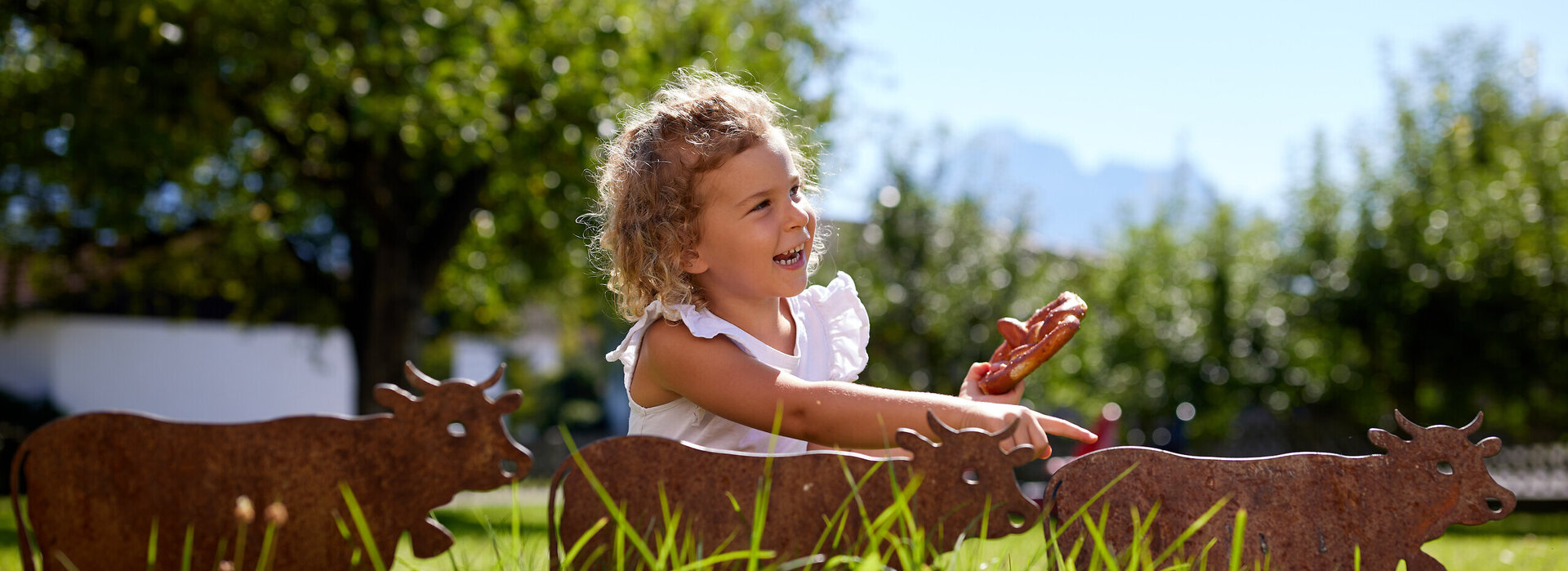 Das Bild zeigt ein Mädchen, das in einer grünen Wiese sitzt und eine Breze in der Hand hat. Im Vordergrund befinden sich 3 kleinere rostige Gartenstecker in Form von Kühen.