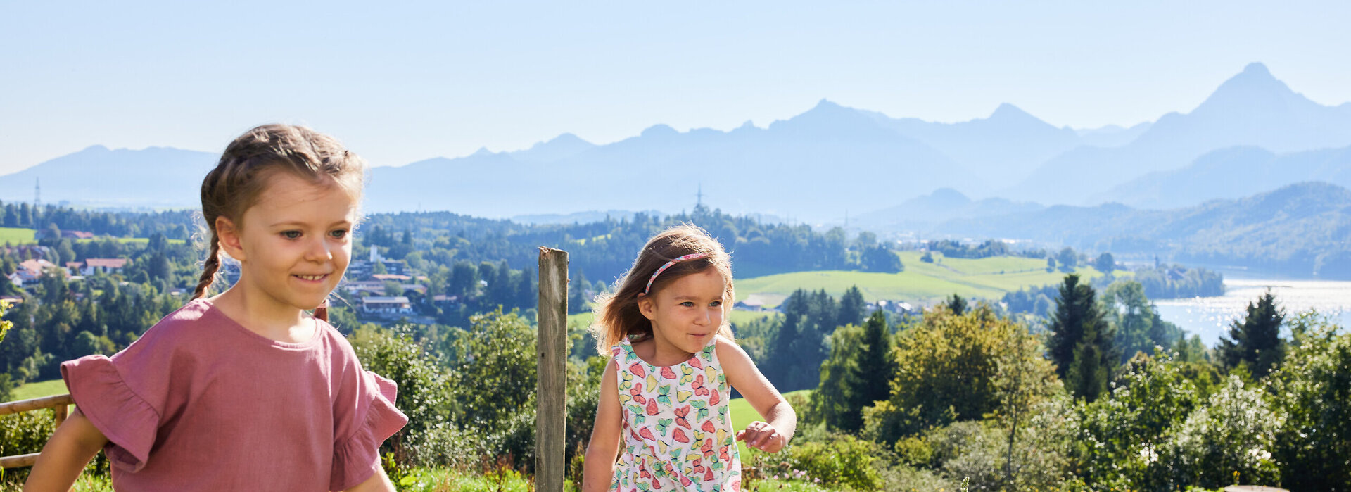 Das Bild fängt einen friedlichen Moment in einem Park ein, wo zwei junge Mädchen einen Spaziergang genießen. Das Mädchen auf der linken Seite, in einem pinkfarbenen Shirt und olivgrünen Shorts gekleidet, marschiert voraus, während das zweite Mädchen, das ein Blumenkleid trägt, dicht hinter ihr herläuft. Sie laufen auf einem Pfad, der von üppigem Grün und Bäumen gesäumt ist. Als Hintergrund des Bildes dient eine atemberaubende Aussicht auf einen See und Berge, was zur Ruhe der Szene beiträgt. Die Mädchen scheinen guter Stimmung zu sein, denn sie beide lächeln. Die Gesamtatmosphäre des Bildes ist friedlich und fröhlich.