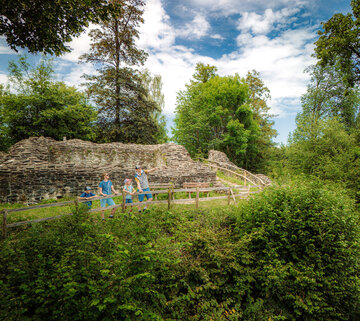 Das Bild zeigt eine Frau, einen Mann und zwei Kinder vor den Mauerresten einer Burg. Die Burg steht auf einer Anhöhe. Die Familie lehnt an einem Holzzaun und blickt ins Tal. Der Mann deutet in die Ferne. Die Burgruine ist umgeben von grünen Laubbäumen. Der Himmel ist blau. Die Sonne scheint.