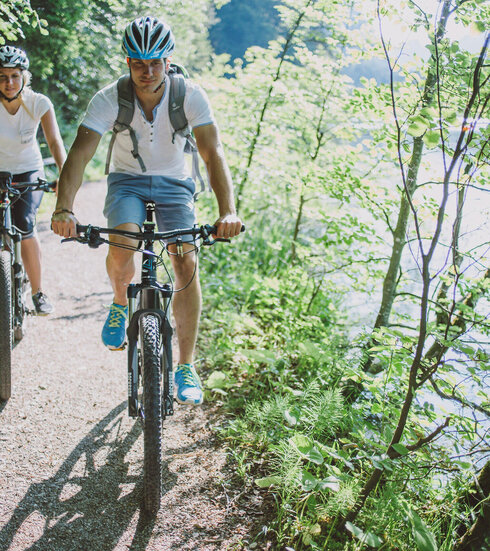 Das Bild zeigt einen Mann und eine Frau beim Fahrradfahren. Sie fahren auf E-Bikes, tragen Freizeitkleidung und Helme. Sie fahren auf den Betrachter zu auf einem unbefestigten Uferweg, der auf der rechten Seite an einen Wald auf der linken Seite an einen See grenzt. Die Sonne scheint.
