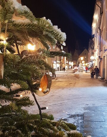 Das Bild fängt eine friedliche Nachtszene in einer Stadt ein. Der Hauptfokus liegt auf einem prächtig geschmückten Weihnachtsbaum, der sich auf der linken Seite des Bildes befindet. Der Baum ist mit weißen Lichtern geschmückt, die der Szene ein festliches Flair verleihen. Der Baum ist von einer schneebedeckten Fläche umgeben, was darauf hinweist, dass es Winter ist.  Im Hintergrund ist eine Straße zu sehen, die von Gebäuden gesäumt wird. Die Straße wird von Straßenlaternen beleuchtet, die auf die Umgebung ein warmes Licht werfen. Einige Menschen können dabei beobachtet werden, wie sie auf der Straße gehen, was ein Gefühl von Leben und Aktivität in die sonst ruhige Szene bringt.  Am Himmel ist es dunkel, was darauf hinweist, dass es Nacht ist. Die allgemeine Atmosphäre des Bildes ist friedlich und festlich und fängt den Charakter einer stillen Winter.