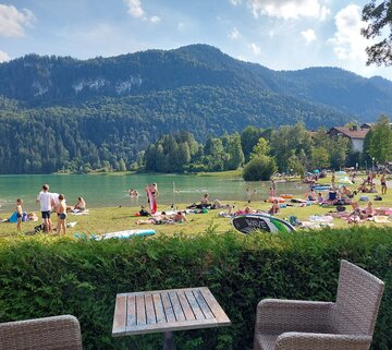 Das Bild zeigt die Liegewiese am Ufer des Badestrandes am Weißensee. Zahlreiche Personen liegen auf bunten Decken und Handtüchern, teilweise unter Sonnenschirmen. Einige sitzen auf Bänken oder stehen am Ufer. Einige Menschen liegen oder stehen im Wasser oder schwimmen. Vereinzelt liegen SUP zwischen den Menschen. Im Vordergrund ist eine hüfthohe, grüne Hecke zu sehen. Davor steht ein Holztisch mit zwei braunen Korbstühlen. Im Hintergrund erhebt sich ein bewaldeter Hügel. Der Himmel ist blau. Die Sonne scheint. 