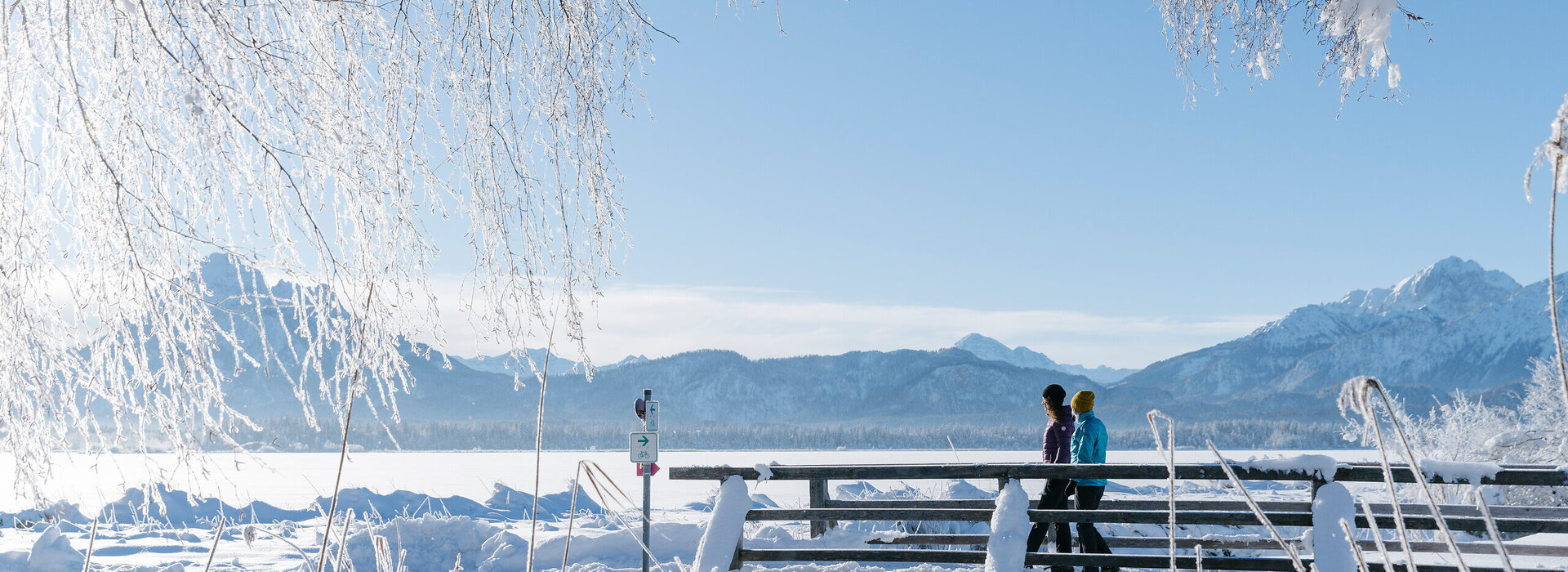 Das Bild zeigt eine verschneite Winterszene. Zwei Personen in warmer Winterkleidung mit Mützen und Handschuhen laufen über eine schneebedeckte Brücke. Die Landschaft um sie herum ist tief verschneit. Der Himmel ist blau. Die Sonne scheint.  
