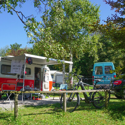 Das Bild zeigt eine friedliche Campingszene. Vorn ist ein rotes und weißes Wohnmobil auf einer grünen Fläche geparkt. Das Wohnmobil ist mit einem Fahrradträger ausgestattet, und neben ihm steht ein Fahrrad. Das Fahrrad hat einen schwarzen Rahmen und rote Akzente.  Im Hintergrund ist ein blaues Wohnmobil auf dem Gras geparkt. Das Wohnmobil ist kleiner als das rote und weiße.  Die Campingplatz ist von Bäumen umgeben, was eine natürliche und friedliche Kulisse schafft. Der Himmel darüber ist klar und blau, was auf einen sonnigen Tag hindeutet. Die Atmosphäre des Bildes ist insgesamt eine des Entspannens und Genießens der Natur.