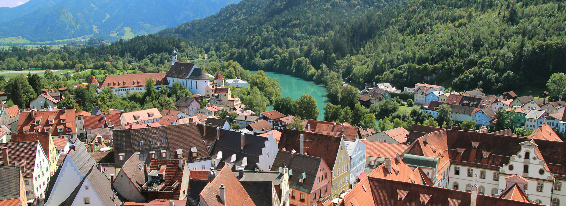 Das Foto zeigt Füssen von oben. Die verwinkelten roten Dächer der Altstadt mit Ihren bunten Fassaden reihen sich aneinander. Im Hintergrund erheben sich von grünem Bergwald bedeckte Berge. Davor fließt der türkisblaue Fluß Lech. 