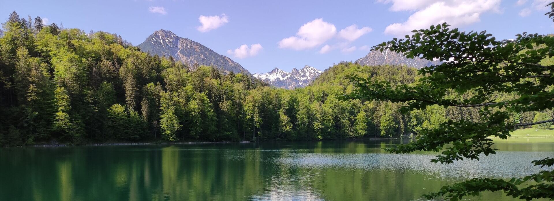 Das Bild fängt eine friedliche Seenlandschaft ein. Der Alatsee ist zu sehen. Der intensiv blaue Alatsee liegt eingebettet in einen üppigen Wald. Die Oberfläche des Wassers ist ruhig, sie spiegelt die umgebende Vegetation und den klaren blauen Himmel darüber wider. Der Himmel ist mit flauschigen weißen Wolken übersät, die zur Ruhe der Szene beitragen. In der Ferne ragen majestätische Berge gegen den Himmel, ihre Gipfel werden mit Schnee bedeckt. 