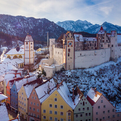 malerisch in einer hauschdünnen Schneedecke, erstreckt sich das Hohe Schloss vor Bergen hervor. Auf dem Bild sieht man die bunten Altstadthäuser von Füssen mit schneebedeckten Dächern. Dahinter das Kloster St. Mang und das Hohe Schloss