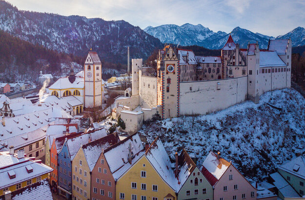 malerisch in einer hauschdünnen Schneedecke, erstreckt sich das Hohe Schloss vor Bergen hervor. Auf dem Bild sieht man die bunten Altstadthäuser von Füssen mit schneebedeckten Dächern. Dahinter das Kloster St. Mang und das Hohe Schloss