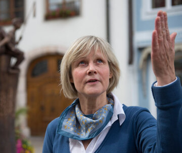Das Bild zeigt eine Gästeführerin mit blondem Haar, die ein blaues Oberteil und ein Schlaal trägt. Sie steht vor einem Brunnen in der Altstadt und zeigt mit der Hand auf etwas, was Sie genauer erklärt. 