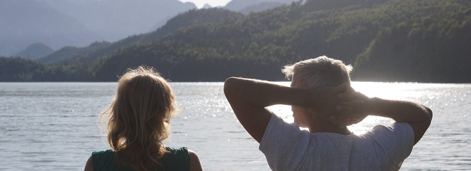 Das Bild zeigt einen Mann und eine Frau, die mit dem Rücken zum Betrachter auf einer Bank sitzen und in die Ferne blicken. Der Mann hat die Arme entspannt hinter dem Kopf verschränkt. Im Hintergrund glitzert der Weißensee im Sonnenlicht und in der Ferne erhebt sich die markante Bergsilhouette des Säulings.