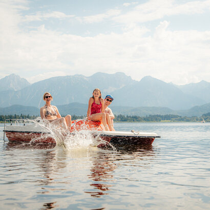 Das Bild zeigt eine idyllische Szene auf einem See, mit drei Personen, die eine Bootsfahrt genießen. Das Boot ist in einem lebhaften Rot lackiert und verfügt über ein weißes Dach, das Schutz für die Passagiere bietet. Das Boot bewegt sich, was durch die aufgewühlten Wasser hinweist, dass es mit einer mäßigen Geschwindigkeit unterwegs ist.  Die drei Personen auf dem Boot sind alle mit Badeanzügen bekleidet, was darauf hinweist, dass sie bereit sind, im See zu schwimmen. Die Person auf der linken Seite sitzt, während die anderen beiden stehen, alle Gesichter zur Kamera gedreht, deren Lächeln Freude und Entspannung ausstrahlen.  Die Kulisse des Bildes ist ein atemberaubender Blick auf Berge, was ein Gefühl von Abenteuer und Entdeckung zur Szene hinzufügt. Der Himmel darüber ist ein klares Blau, mit flauschigen weißen Wolken, die das malerische Setting vervollständigen. Das Gesamtbild erzeugt ein Bild eines perfekten Tages am Wasser, umgeben von der Schönheit der Natur.