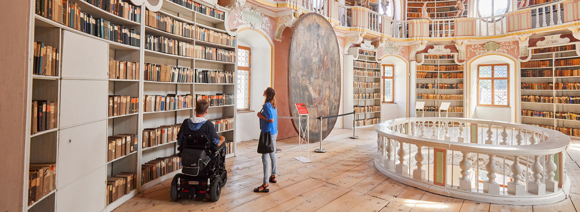 Die Aufnahme dokumentiert eine friedliche Szene in einer Bibliothek. Die Bibliothek wird durch ihre hohe Decke und den Holzboden geprägt, was ihr eine warme und einladende Atmosphäre verleiht. Die Wände sind mit Bücherregalen geschmückt, die eine Vielzahl von Büchern aufweisen und auf eine umfangreiche Sammlung von Wissen hinweisen.  In der Mitte des Raumes sitzt ein Mann im Rollstuhl und ist vertieft in das Lesen eines Buches. Seine Position deutet darauf hin, dass er sich in dieser Umgebung wohl und entspannt fühlt. In der Nähe ist ein junges Mädchen zu sehen, das nach dem Mann im Rollstuhl schaut. Ihre Neugier ist deutlich erkennbar, während sie ihn beobachtet.  Der Raum wird weiter durch ein großes rundes Fenster aufgewertet, das natürliches Licht hereinlässt und das Licht auf die Bücher fällt, wodurch eine friedliche Atmosphäre geschaffen wird. Das Gesamtbild zeichnet ein Bild eines ruhigen Nachmittags, der in der Beschäftigung mit Wissen verbracht wird.