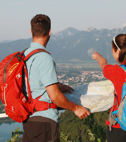 Das Bild fängt einen Moment der Abenteuer und Erkundung ein. Zwei Personen, einem Mann und einer Frau, stehen auf einem Berg, ihre Rücken zeigen zur Kamera. Sie sind beide mit Rucksäcken ausgestattet, was darauf hinweist, dass sie auf einem Hike oder Trek sind. Der Mann deutet auf eine Karte. Die Frau deutet in die Ferne. Der Hintergrund sieht man über eine Ebene aus Dörfern, See und Natur. Der Himmel über ihnen ist blau und klar.