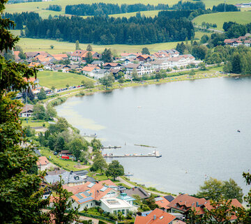 Im Fokus des Bildes steht ein See, um den zahlreiche Häuser stehen. Das Foto wurde von einer Anhöhe aus aufgenommen. Die Häuser haben rote Dächer und weiße Fassaden. An das Ufer grenzt eine Wiese, die durch einen befestigten, rundumführenden Fußweg unterbrochen wird. Auf der Uferwiese sind Badegäste zu sehen. Im Wasser schwimmen vereinzelte Boote.  