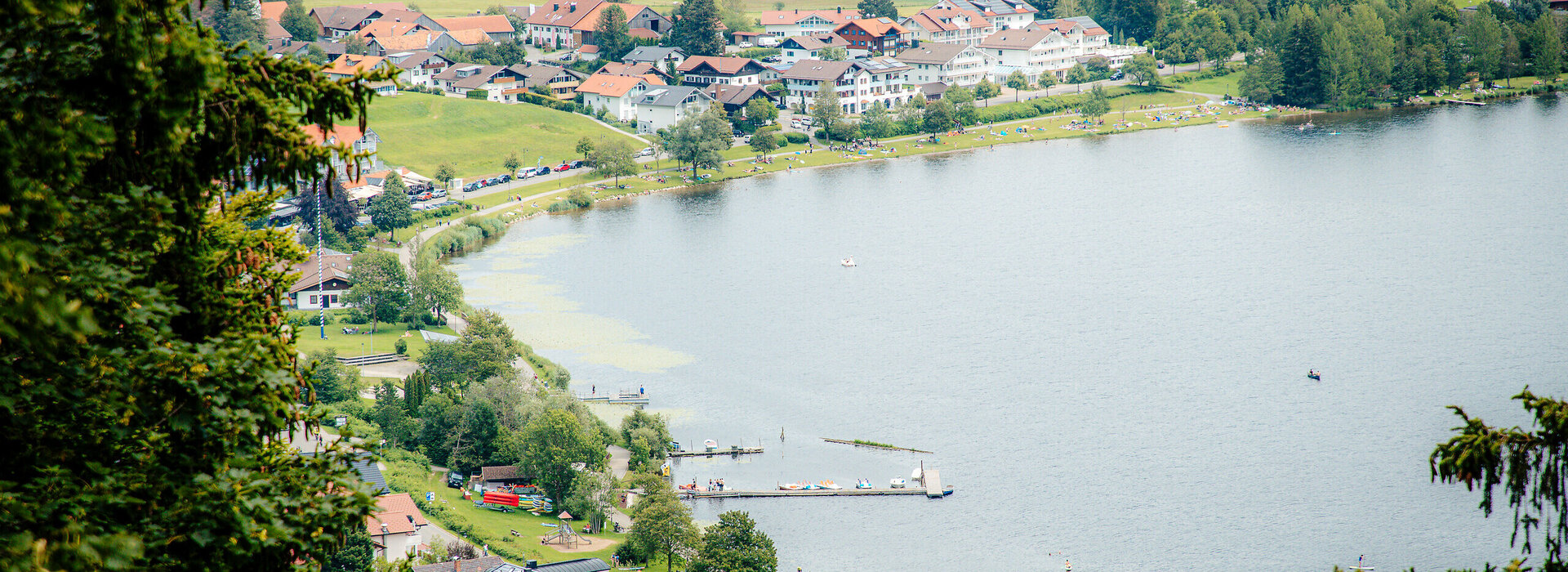 Im Fokus des Bildes steht ein See, um den zahlreiche Häuser stehen. Das Foto wurde von einer Anhöhe aus aufgenommen. Die Häuser haben rote Dächer und weiße Fassaden. An das Ufer grenzt eine Wiese, die durch einen befestigten, rundumführenden Fußweg unterbrochen wird. Auf der Uferwiese sind Badegäste zu sehen. Im Wasser schwimmen vereinzelte Boote.  
