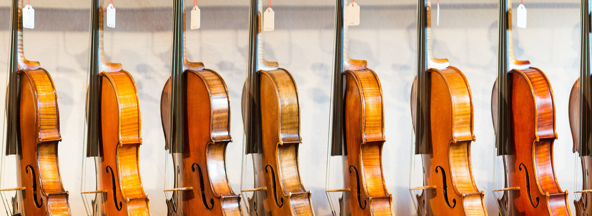 Das Bild zeigt eine Sammlung von Geigen, die jeder einzeln von einem Haken an einer Wand hängt. Die Geigen sind in einer Reihe angeordnet, mit jedem Instrument gleich in eine Richtung zeigend. Die Geigen sind unterschiedlicher Größe, was darauf hindeutet, dass sie für verschiedene Altersgruppen oder Spielweisen konzipiert sind. Die Geigen sind aus Holz und haben eine reiche, warme Braunfärbung, die vor dem weißen Hintergrund im Bild gut zur Geltung kommt. Die Saiten der Geigen sind sichtbar und tragen dazu bei, die Gesamtästhetik des Bildes zu vervollständigen. Die Anordnung der Geigen und deren Positionierung an der Wand erzeugen ein Gefühl von Ordnung und Harmonie