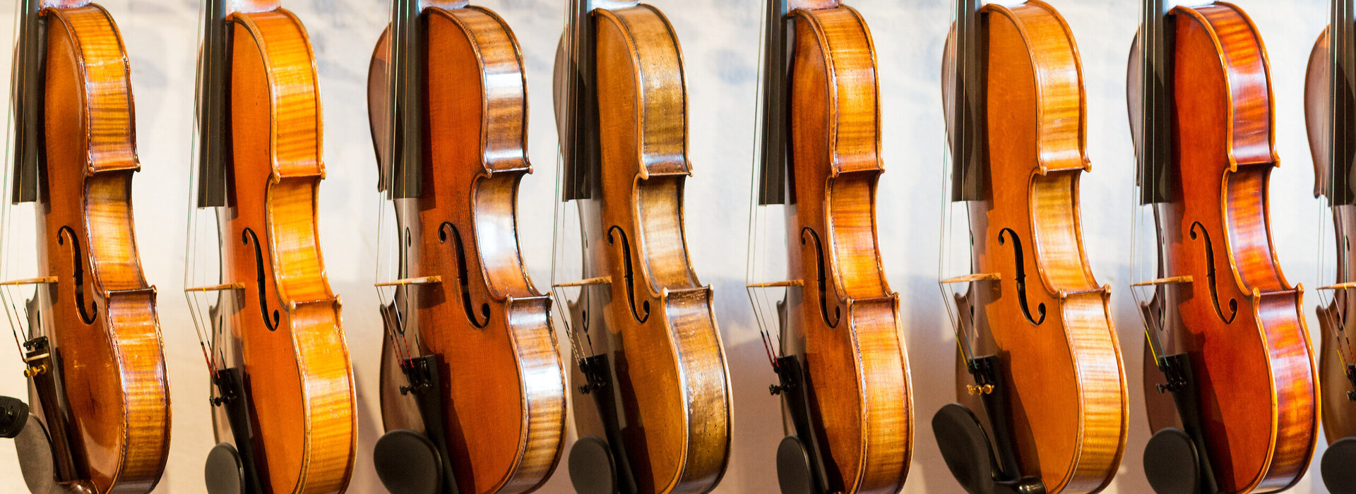 Das Bild zeigt eine Sammlung von Geigen, die jeder einzeln von einem Haken an einer Wand hängt. Die Geigen sind in einer Reihe angeordnet, mit jedem Instrument gleich in eine Richtung zeigend. Die Geigen sind unterschiedlicher Größe, was darauf hindeutet, dass sie für verschiedene Altersgruppen oder Spielweisen konzipiert sind. Die Geigen sind aus Holz und haben eine reiche, warme Braunfärbung, die vor dem weißen Hintergrund im Bild gut zur Geltung kommt. Die Saiten der Geigen sind sichtbar und tragen dazu bei, die Gesamtästhetik des Bildes zu vervollständigen. Die Anordnung der Geigen und deren Positionierung an der Wand erzeugen ein Gefühl von Ordnung und Harmonie