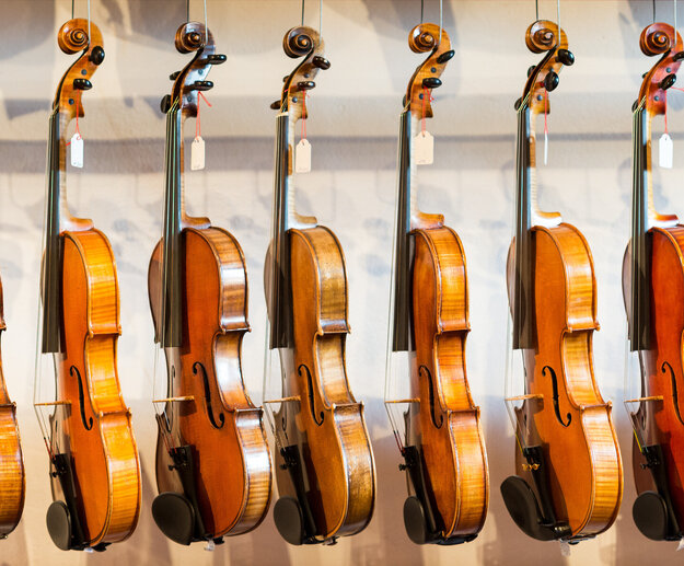 Das Bild zeigt eine Sammlung von Geigen, die jeder einzeln von einem Haken an einer Wand hängt. Die Geigen sind in einer Reihe angeordnet, mit jedem Instrument gleich in eine Richtung zeigend. Die Geigen sind unterschiedlicher Größe, was darauf hindeutet, dass sie für verschiedene Altersgruppen oder Spielweisen konzipiert sind. Die Geigen sind aus Holz und haben eine reiche, warme Braunfärbung, die vor dem weißen Hintergrund im Bild gut zur Geltung kommt. Die Saiten der Geigen sind sichtbar und tragen dazu bei, die Gesamtästhetik des Bildes zu vervollständigen. Die Anordnung der Geigen und deren Positionierung an der Wand erzeugen ein Gefühl von Ordnung und Harmonie