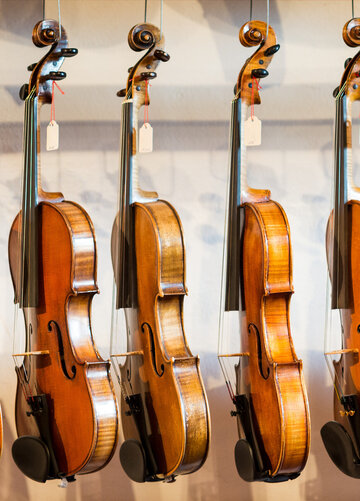 Das Bild zeigt eine Sammlung von Geigen, die jeder einzeln von einem Haken an einer Wand hängt. Die Geigen sind in einer Reihe angeordnet, mit jedem Instrument gleich in eine Richtung zeigend. Die Geigen sind unterschiedlicher Größe, was darauf hindeutet, dass sie für verschiedene Altersgruppen oder Spielweisen konzipiert sind. Die Geigen sind aus Holz und haben eine reiche, warme Braunfärbung, die vor dem weißen Hintergrund im Bild gut zur Geltung kommt. Die Saiten der Geigen sind sichtbar und tragen dazu bei, die Gesamtästhetik des Bildes zu vervollständigen. Die Anordnung der Geigen und deren Positionierung an der Wand erzeugen ein Gefühl von Ordnung und Harmonie