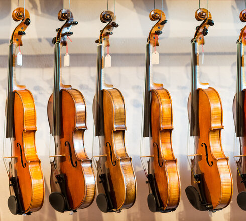 Das Bild zeigt eine Sammlung von Geigen, die jeder einzeln von einem Haken an einer Wand hängt. Die Geigen sind in einer Reihe angeordnet, mit jedem Instrument gleich in eine Richtung zeigend. Die Geigen sind unterschiedlicher Größe, was darauf hindeutet, dass sie für verschiedene Altersgruppen oder Spielweisen konzipiert sind. Die Geigen sind aus Holz und haben eine reiche, warme Braunfärbung, die vor dem weißen Hintergrund im Bild gut zur Geltung kommt. Die Saiten der Geigen sind sichtbar und tragen dazu bei, die Gesamtästhetik des Bildes zu vervollständigen. Die Anordnung der Geigen und deren Positionierung an der Wand erzeugen ein Gefühl von Ordnung und Harmonie